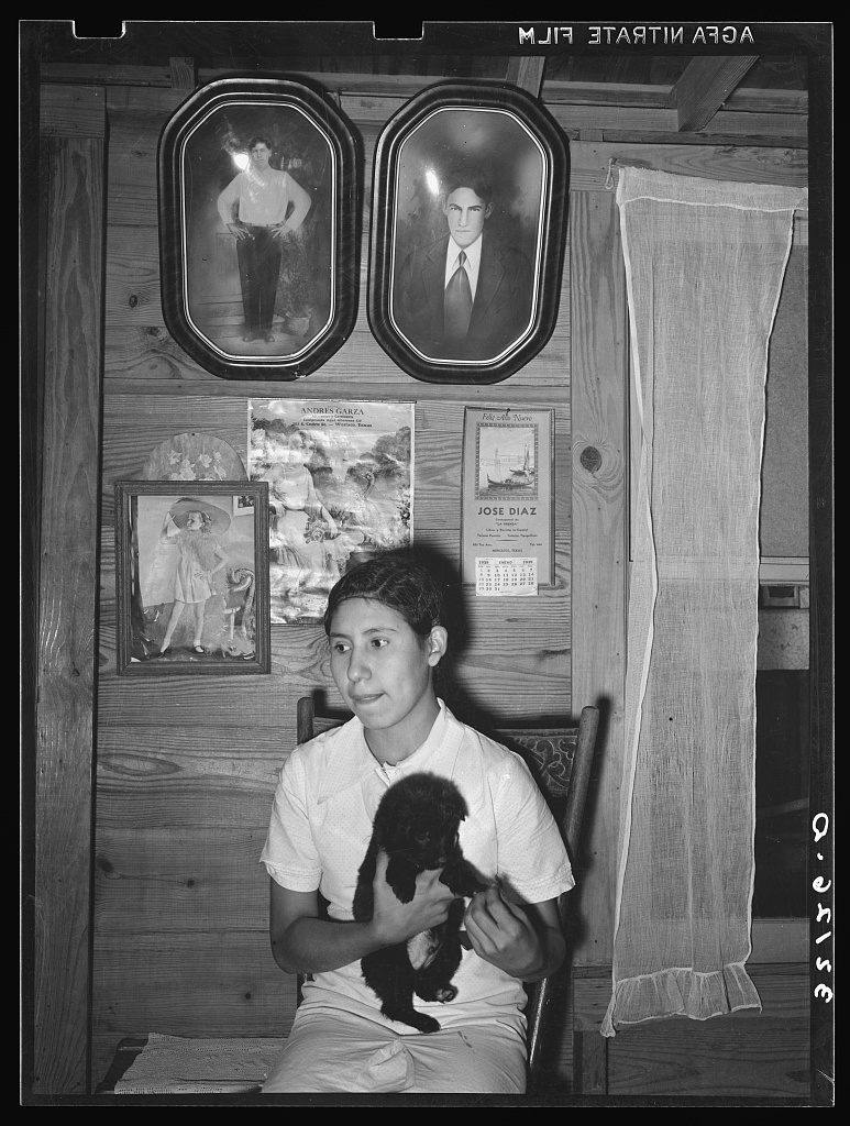 Daughter of Mexican farm family at home. Portraits of family are above her. Near Santa Maria, Texas, 1939 (Library of Congress)