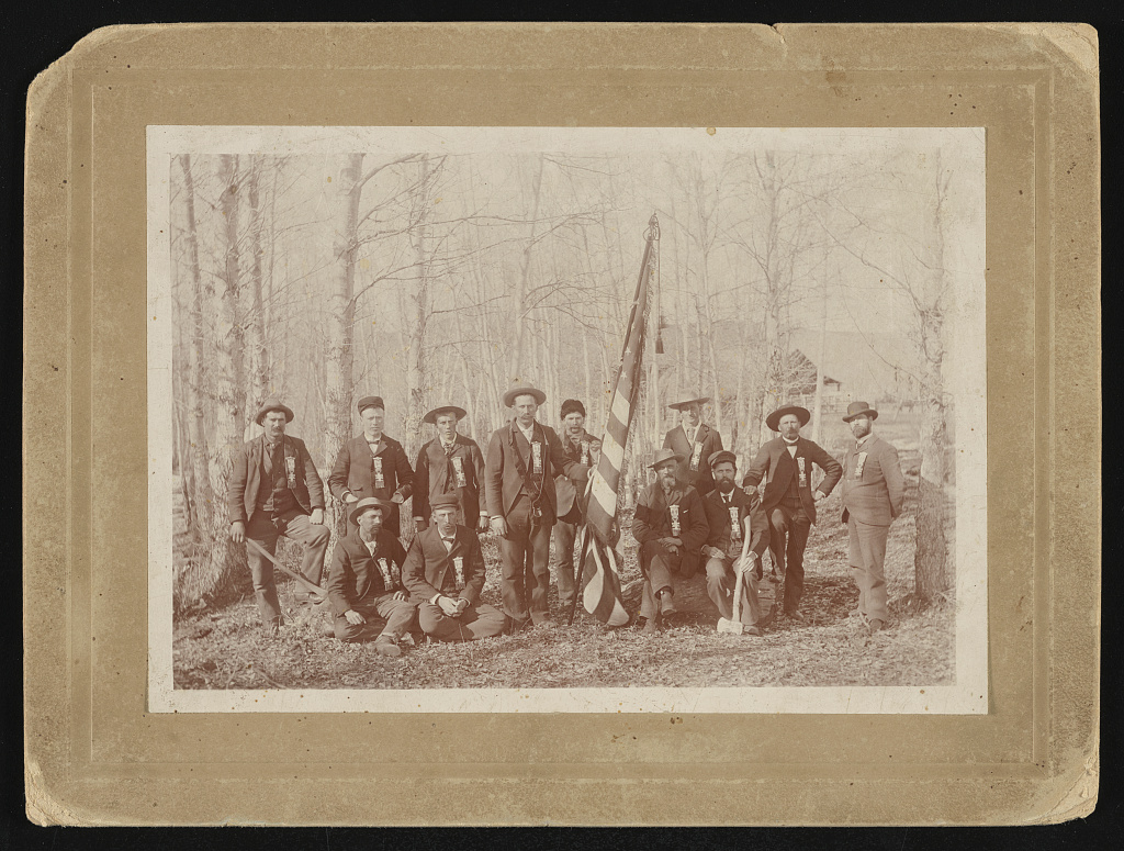 [Group of Civil War veterans outdoors, wearing large ribbon badges, some holding axes and an American flag, possibly at a reunion for the Third Battle of Winchester, Virginia], 1880 (Library of Congress)